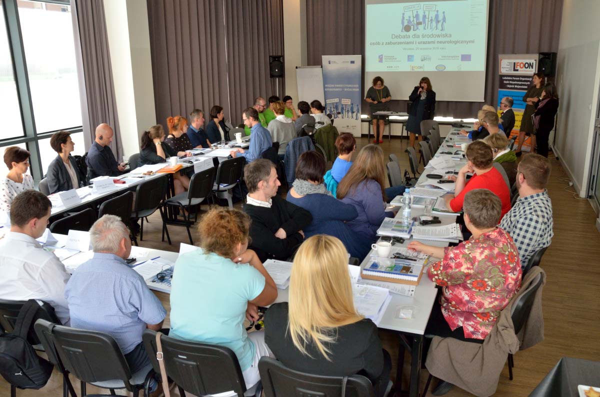 Początek debaty. Widok na salę konferencyjną w kierunku ekranu multimedialnego. Liczni uczestnicy usadzeni przy podkowie.