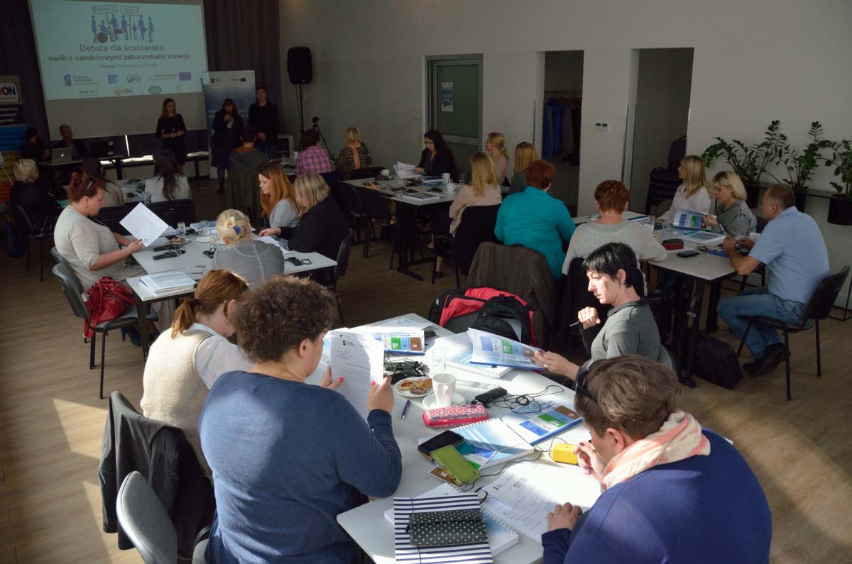 Początek debaty. Widok na salę konferencyjną zza wysp roboczych. W tle ekran multimedialny i moderatorzy debat. Uczestnicy siedzący przy wyspach roboczych na pierwszym planie.