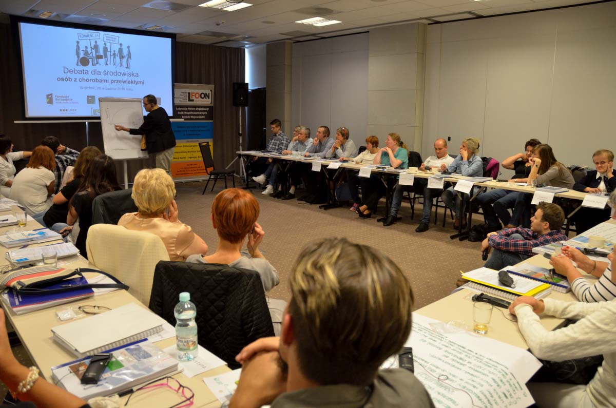 Widok na salę konferencyjną. Uczestnicy debaty ustawieni w&nbsp;podkowie. Na dalszym planie Pan Wubbe rysujący wykres na flipczarcie.