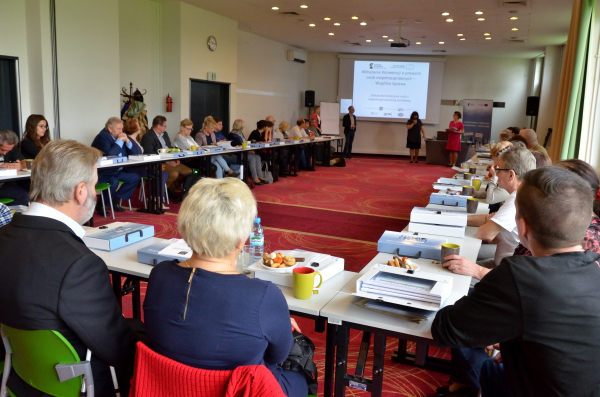 Początek debaty. Widok z rogu sali konferencyjnej. Uczestnicy usadzeni w podkowie. Prowadzący stoją przodem do uczestników.