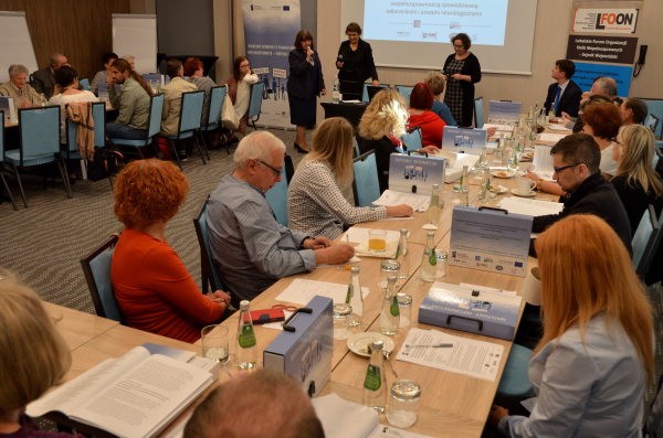 Widok na salę konferencyjną z rogu sali. Początek debaty. Prowadzący stoją przodem do uczestników.