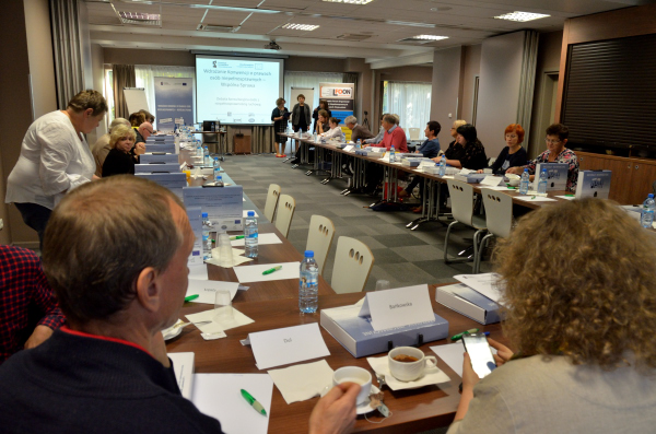 Widok na salę konferencyjną z lewego rogu sali. Uczestnicy usadzeni w podkowie. Początek debaty. Prowadzący stoją przed ekranem przodem do uczestników.